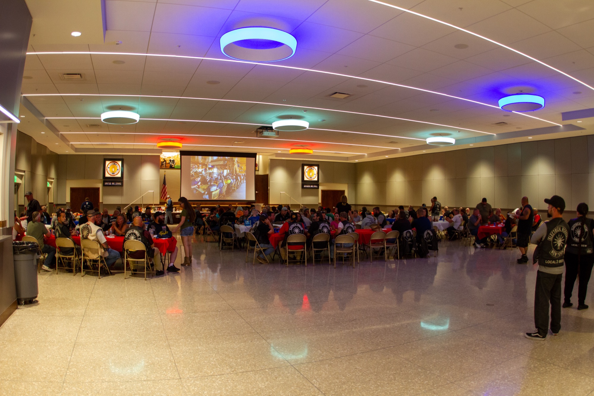 A packed evening event inside the IBEW Local 1 hall, tables set under colored lights with a presentation on the big screen.