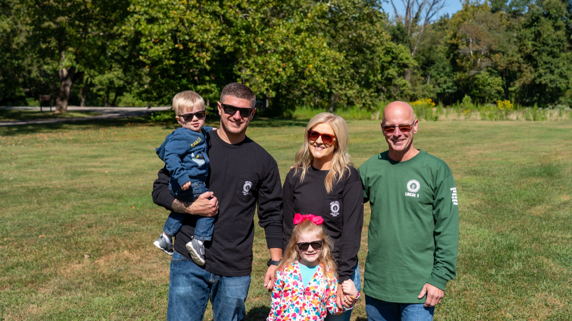Greg Booth's daughter and her family standing in a sunlit grassy area in EWRC1 club shirts, with IBEW Local 1 Representative Jeff Witt on the far right.